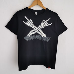 May include: A black short-sleeve t-shirt featuring a graphic of two white skeleton arms crossed in a rock-and-roll hand gesture. Below the arms is Japanese text in a grey, angular font. A small red circular logo is visible at the bottom hem.