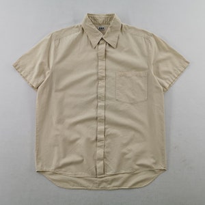 May include: A short-sleeved button-down shirt in a solid beige color. The shirt features a standard collar, a front button placket, and a single chest pocket on the left side. A small white label with the text A.P.C. is visible at the inner neckline.