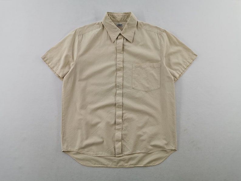 May include: A short-sleeved button-down shirt in a solid beige color. The shirt features a standard collar, a front button placket, and a single chest pocket on the left side. A small white label with the text A.P.C. is visible at the inner neckline.