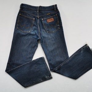 May include: Dark wash denim jeans with a flared leg. The jeans have a classic five-pocket design, a brown leather patch with the "Wrangler" logo, and the signature "W" stitching on the back pockets. The jeans are laid flat on a white surface.