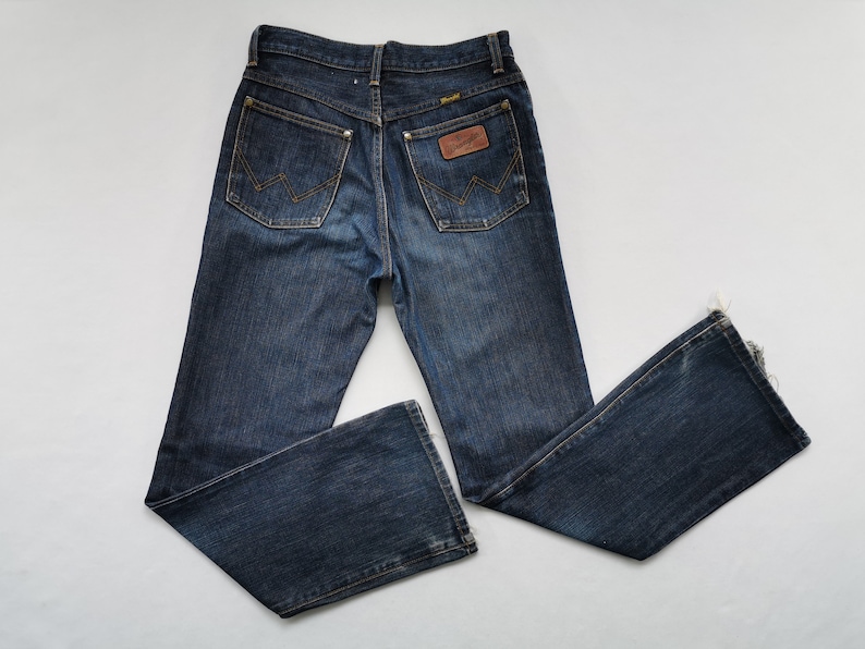 May include: Dark wash denim jeans with a flared leg. The jeans have a classic five-pocket design, a brown leather patch with the "Wrangler" logo, and the signature "W" stitching on the back pockets. The jeans are laid flat on a white surface.