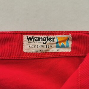 May include: Close-up of red Wrangler jeans. The label reads "Wrangler JEANS & SPORTSWEAR SIZE 34 INS / 86 CM". The label also includes Japanese text. The jeans are a vibrant red color.