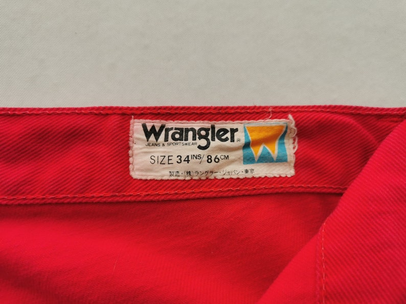 May include: Close-up of red Wrangler jeans. The label reads "Wrangler JEANS & SPORTSWEAR SIZE 34 INS / 86 CM". The label also includes Japanese text. The jeans are a vibrant red color.