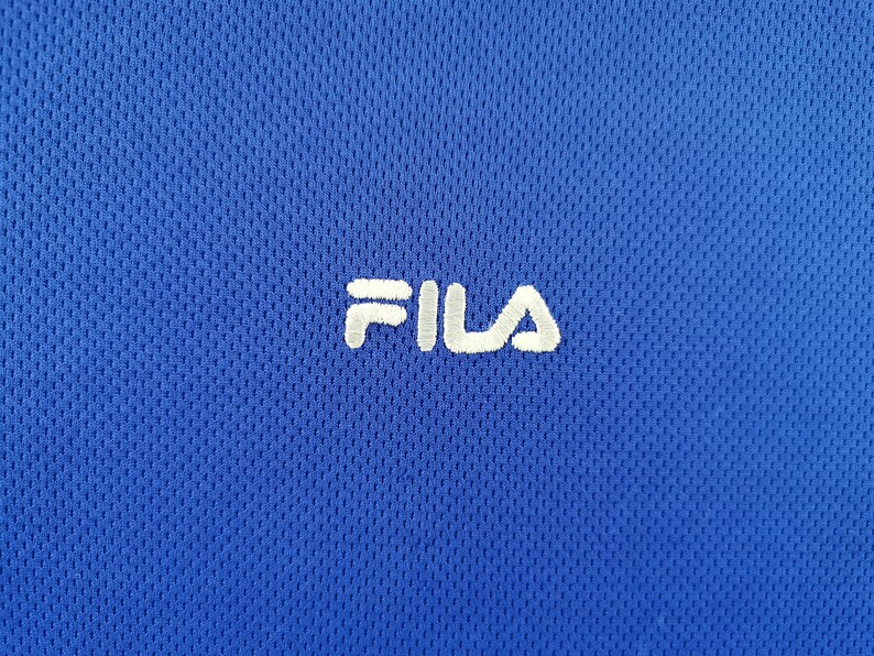 Puede incluir: Primer plano de una camiseta deportiva azul real con tejido texturizado y transpirable. El logotipo blanco "FILA" bordado est&aacute; centrado en el pecho. El dise&ntilde;o de la camiseta sugiere que est&aacute; hecha para deportes o uso casual.