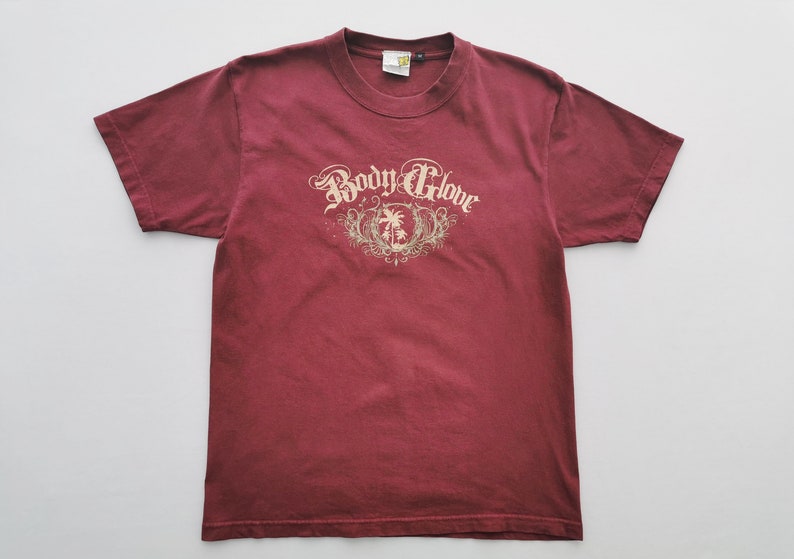 May include: A maroon t-shirt with a crew neck and short sleeves. The front displays the text "Body Glove" in an ornate font above a palm tree design. The shirt is made of a soft material and is laid flat on a white surface.