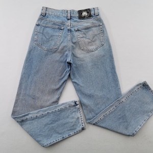 May include: Light blue denim jeans with a faded wash. The jeans have a classic five-pocket design, a straight leg, and a black leather patch with a logo on the back. The jeans are laid flat.
