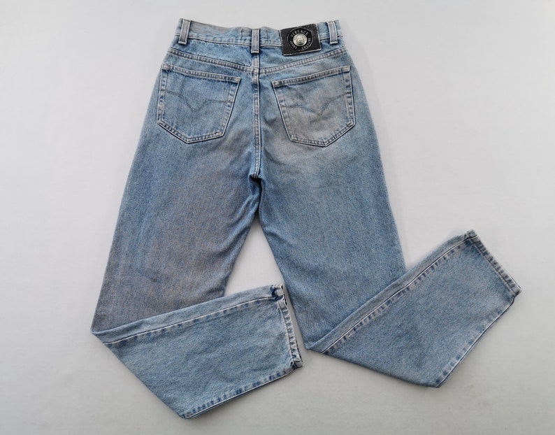 May include: Light blue denim jeans with a faded wash. The jeans have a classic five-pocket design, a straight leg, and a black leather patch with a logo on the back. The jeans are laid flat.