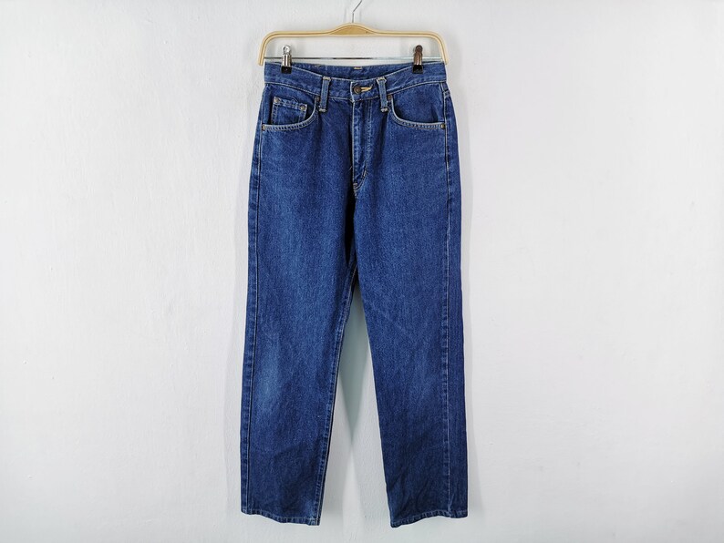 Puede incluir: Un par de vaqueros cl&aacute;sicos azules colgados de una percha de madera. Los vaqueros tienen un corte recto, dise&ntilde;o de cinco bolsillos y cierre de bot&oacute;n y cremallera. El denim tiene un lavado medio con sutiles decoloraciones.