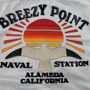 May include: White shirt with an embroidered design featuring a sunset, seagull, and text. The text reads "BREEZY POINT NAVAL STATION ALAMEDA CALIFORNIA" in black. The sunset is in shades of red, orange, and yellow.