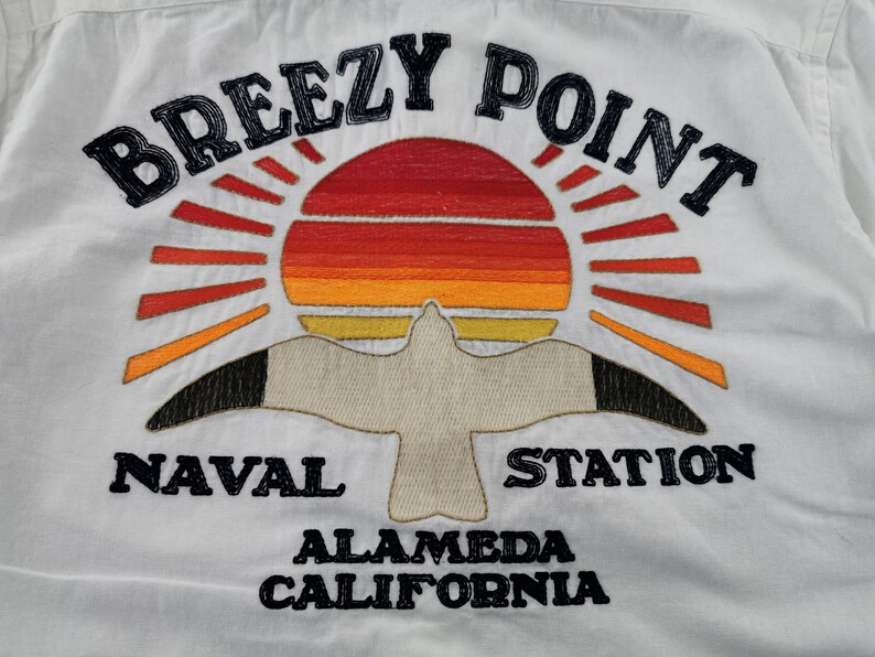 May include: White shirt with an embroidered design featuring a sunset, seagull, and text. The text reads "BREEZY POINT NAVAL STATION ALAMEDA CALIFORNIA" in black. The sunset is in shades of red, orange, and yellow.