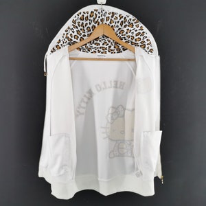 May include: White zip-up hoodie with a leopard print lining in the hood. The hoodie features a Hello Kitty graphic on the front and the words "Hello Kitty" printed in a mirrored design. The hoodie has two front pockets and is hanging on a wooden hanger.