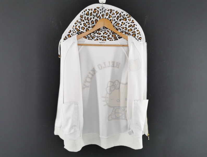 May include: White zip-up hoodie with a leopard print lining in the hood. The hoodie features a Hello Kitty graphic on the front and the words "Hello Kitty" printed in a mirrored design. The hoodie has two front pockets and is hanging on a wooden hanger.