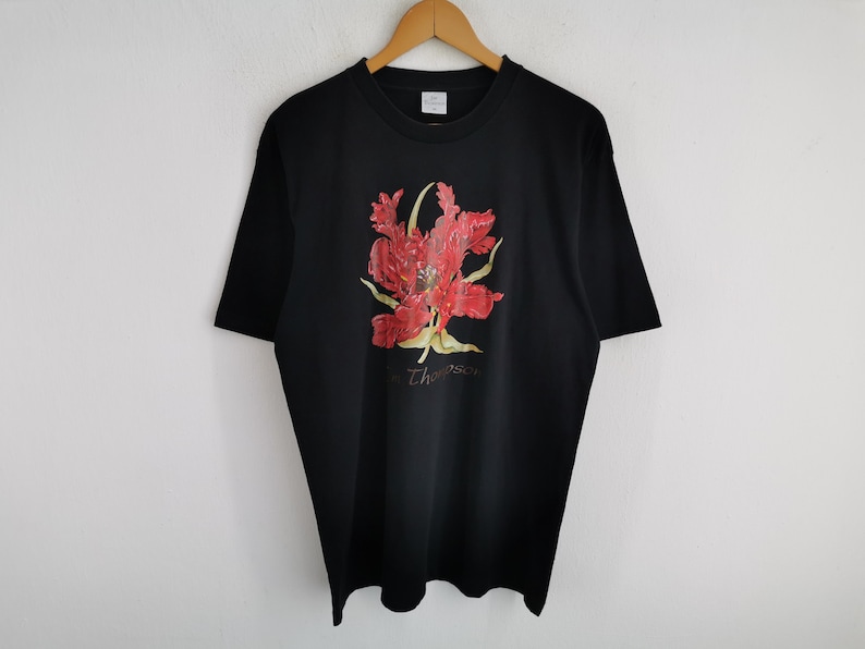 May include: Black t-shirt featuring a vibrant red and yellow floral design. The design includes the name "Thompson" in a cursive font. The shirt is hanging on a wooden hanger against a white background.