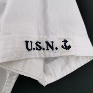 May include: Close-up of a white short-sleeved shirt. The cuff features the embroidered letters "U.S.N." in navy blue, along with a small anchor symbol. The shirt is made of a light, possibly linen, fabric.