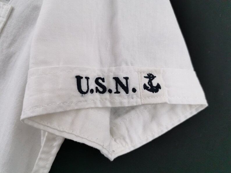 May include: Close-up of a white short-sleeved shirt. The cuff features the embroidered letters "U.S.N." in navy blue, along with a small anchor symbol. The shirt is made of a light, possibly linen, fabric.