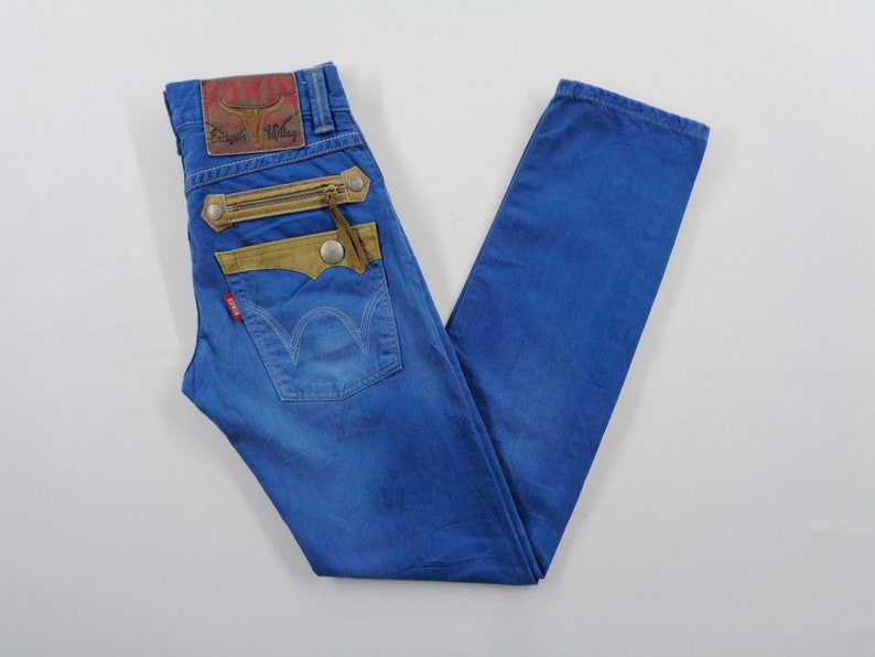 K&ouml;nnte beinhalten: Blaue Jeans aus Denim mit einzigartigen Designelementen. Mit einem braunen Lederaufn&auml;her, einer Rei&szlig;verschlusstasche und einem roten Logo-Etikett. Die Jeans haben ein klassisches F&uuml;nf-Taschen-Design und einen geraden Beinverlauf.
