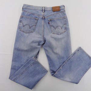 May include: Light blue denim jeans with a classic five-pocket design. The jeans feature a faded wash, a brown leather patch on the back, and a red tag on the back pocket. The jeans are laid flat on a white surface.