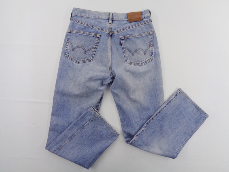 May include: Light blue denim jeans with a classic five-pocket design. The jeans feature a faded wash, a brown leather patch on the back, and a red tag on the back pocket. The jeans are laid flat on a white surface.