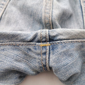 Puede incluir: Primer plano de unos vaqueros de mezclilla azul claro. La imagen destaca los detalles de las costuras, con costuras visibles y detalles de hilo amarillo. La tela tiene un aspecto texturizado, mostrando el tejido de la mezclilla.