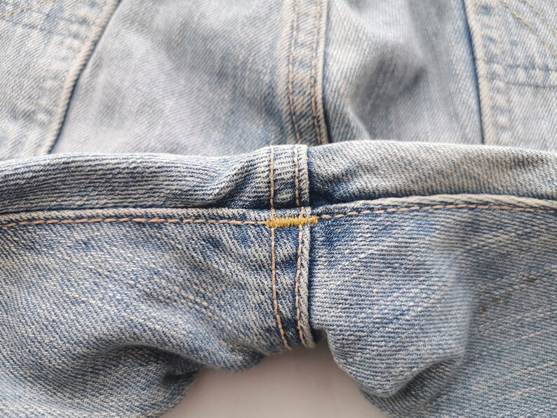 Puede incluir: Primer plano de unos vaqueros de mezclilla azul claro. La imagen destaca los detalles de las costuras, con costuras visibles y detalles de hilo amarillo. La tela tiene un aspecto texturizado, mostrando el tejido de la mezclilla.