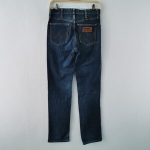 May include: Dark blue denim jeans with a classic straight-leg fit. Features include a brown leather patch with the "Wrangler" logo and signature stitching on the back pockets. The jeans are hanging on a metal hanger.