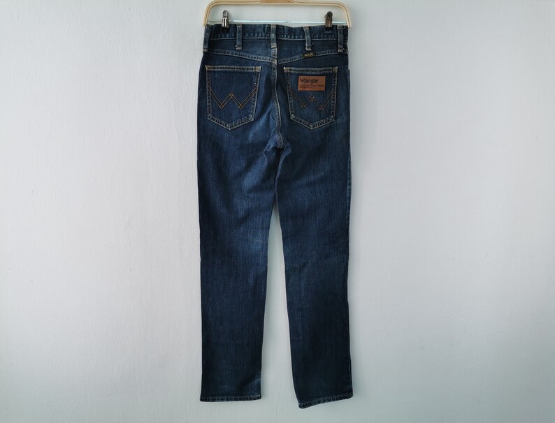 May include: Dark blue denim jeans with a classic straight-leg fit. Features include a brown leather patch with the "Wrangler" logo and signature stitching on the back pockets. The jeans are hanging on a metal hanger.