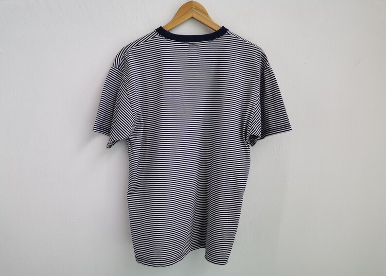 Puede incluir: Camiseta a rayas azul marino y blanca de manga corta, colgada de una percha de madera. La camiseta tiene cuello redondo azul marino. Las rayas horizontales son uniformes en todo el tejido.