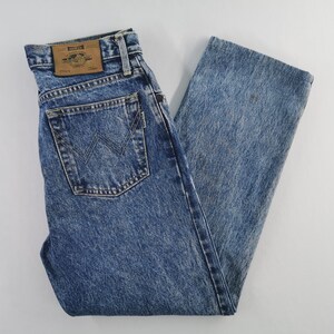 Puede incluir: Vaqueros azules con lavado &aacute;cido con un dise&ntilde;o cl&aacute;sico de cinco bolsillos. Los vaqueros presentan un logotipo de Wrangler en el bolsillo trasero y un parche de cuero en la cintura. El denim tiene un aspecto descolorido y desgastado.