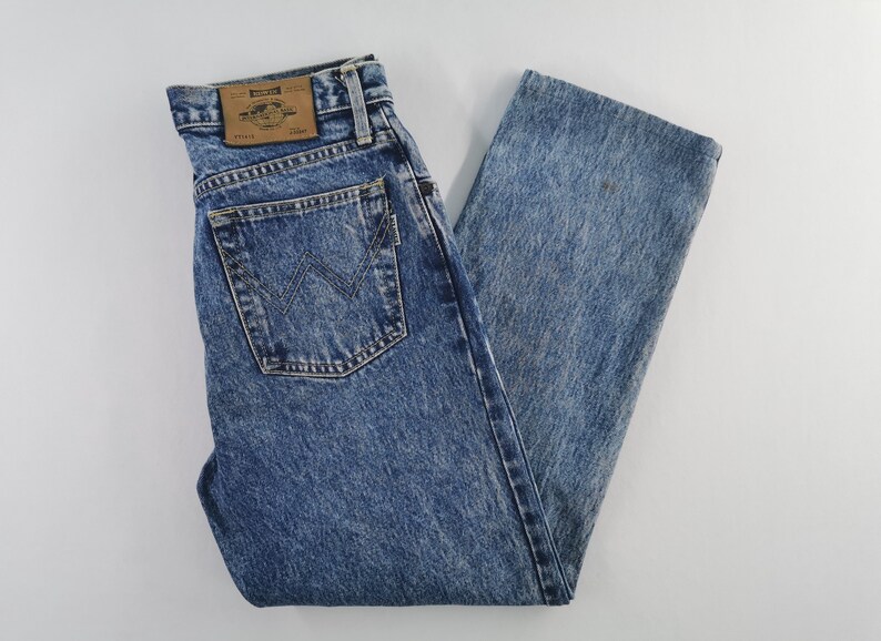 Puede incluir: Vaqueros azules con lavado &aacute;cido con un dise&ntilde;o cl&aacute;sico de cinco bolsillos. Los vaqueros presentan un logotipo de Wrangler en el bolsillo trasero y un parche de cuero en la cintura. El denim tiene un aspecto descolorido y desgastado.