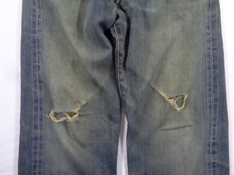 Puede incluir: Vaqueros de mezclilla azul descolorido con detalles desgastados, incluyendo dos roturas a la altura de la rodilla. Los vaqueros tienen un dise&ntilde;o cl&aacute;sico de cinco bolsillos y un lavado claro, mostrando signos de desgaste.