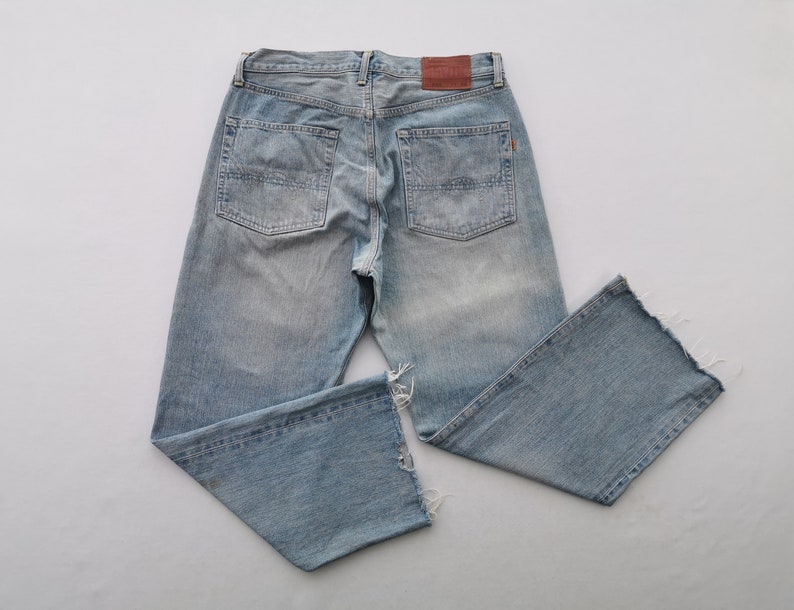Puede incluir: Vaqueros de mezclilla azul claro con un lavado descolorido y detalles desgastados. Los vaqueros presentan un dise&ntilde;o cl&aacute;sico de cinco bolsillos, un parche de cuero marr&oacute;n y dobladillos sin rematar y deshilachados. Los vaqueros est&aacute;n planos sobre un fondo blanco.