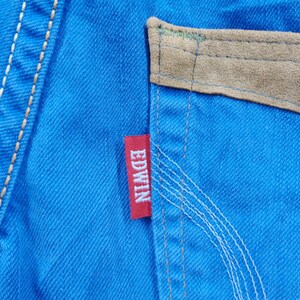 K&ouml;nnte beinhalten: Nahaufnahme von leuchtend blauem Denim-Stoff mit einem Wildleder-Taschendetail in Beige. Ein rotes "EDWIN"-Etikett ist auf den Denim gen&auml;ht. Wei&szlig;e N&auml;hte akzentuieren die N&auml;hte und die Tasche.