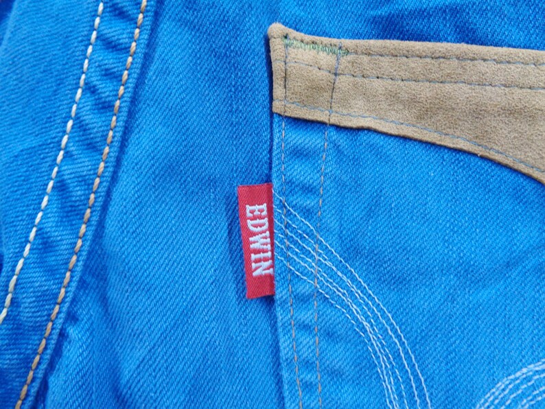 K&ouml;nnte beinhalten: Nahaufnahme von leuchtend blauem Denim-Stoff mit einem Wildleder-Taschendetail in Beige. Ein rotes "EDWIN"-Etikett ist auf den Denim gen&auml;ht. Wei&szlig;e N&auml;hte akzentuieren die N&auml;hte und die Tasche.