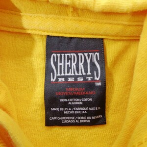 Op de afbeelding: Close-up van een felgele hoodie met een zwart label. Het label toont "SHERRY'S BEST" in witte letters. Het label geeft ook de maat medium en 100% katoen aan. Gemaakt in de VS.