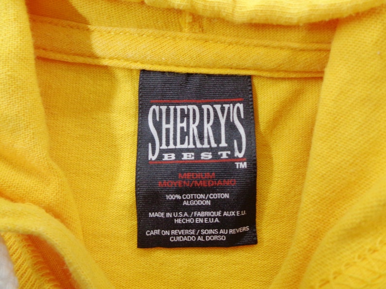 Op de afbeelding: Close-up van een felgele hoodie met een zwart label. Het label toont "SHERRY'S BEST" in witte letters. Het label geeft ook de maat medium en 100% katoen aan. Gemaakt in de VS.