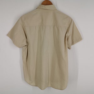 May include: A short-sleeved button-down shirt in a solid beige color. The shirt features a standard collar, a horizontal back yoke with subtle pleating detail, and a curved hemline. The garment is displayed hanging on a wooden hanger.