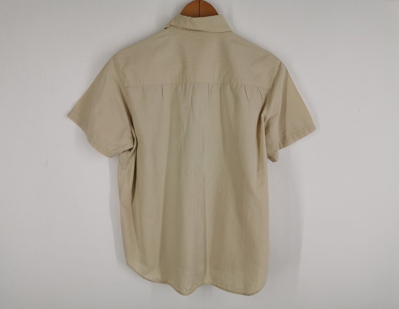 May include: A short-sleeved button-down shirt in a solid beige color. The shirt features a standard collar, a horizontal back yoke with subtle pleating detail, and a curved hemline. The garment is displayed hanging on a wooden hanger.