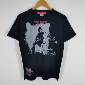 Może przedstawiać: Czarny t-shirt z nadrukiem graficznym przedstawiającym członka zespołu oraz słowa "STRUMMER" i "STREET TONE" w kolorze różowym i białym. Koszulka ma również dodatkowy tekst i małe logo. Koszulka wisi na drewnianym wieszaku.