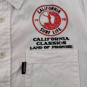 May include: White button-down shirt with a pocket and embroidered design. The design features a red circle with the words "CALIFORNIA SURF LIFE" and a surfer graphic. Below the circle are the words "CALIFORNIA CLASSICS LAND OF PROMISE."