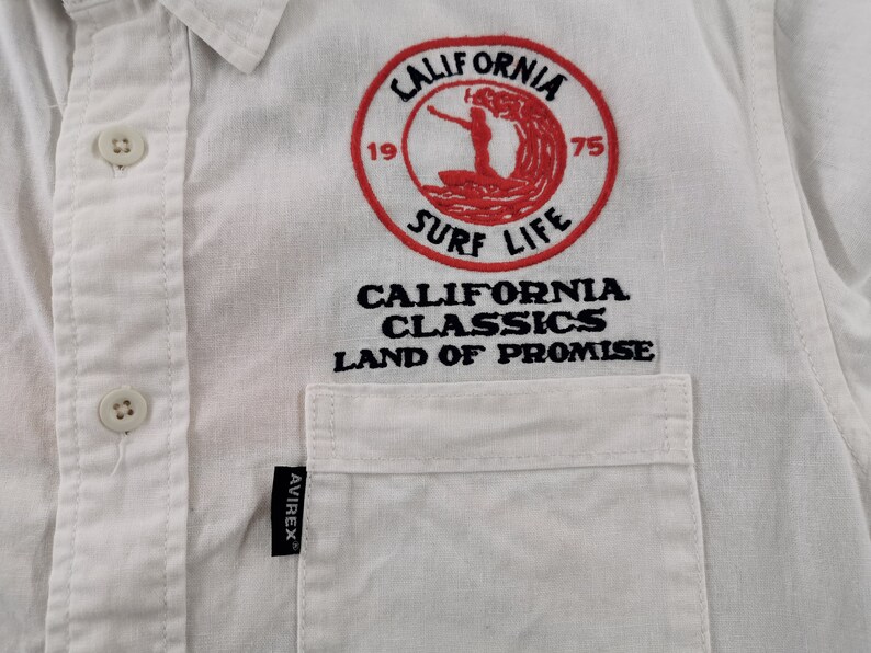 May include: White button-down shirt with a pocket and embroidered design. The design features a red circle with the words "CALIFORNIA SURF LIFE" and a surfer graphic. Below the circle are the words "CALIFORNIA CLASSICS LAND OF PROMISE."