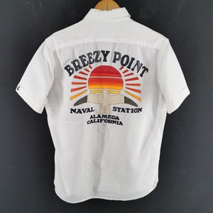 May include: White short-sleeved shirt with a colourful embroidered design on the back. The design features a sunset, a bird, and the words "Breezy Point Naval Station Alameda California". The shirt is hanging on a wooden hanger.