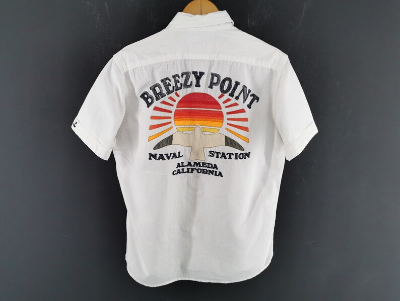 May include: White short-sleeved shirt with a colourful embroidered design on the back. The design features a sunset, a bird, and the words "Breezy Point Naval Station Alameda California". The shirt is hanging on a wooden hanger.