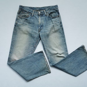 May include: Light blue denim jeans with a faded wash and a distressed knee. The jeans have a classic five-pocket design, a button and zip closure, and a straight leg. The jeans are laid flat on a light blue surface.
