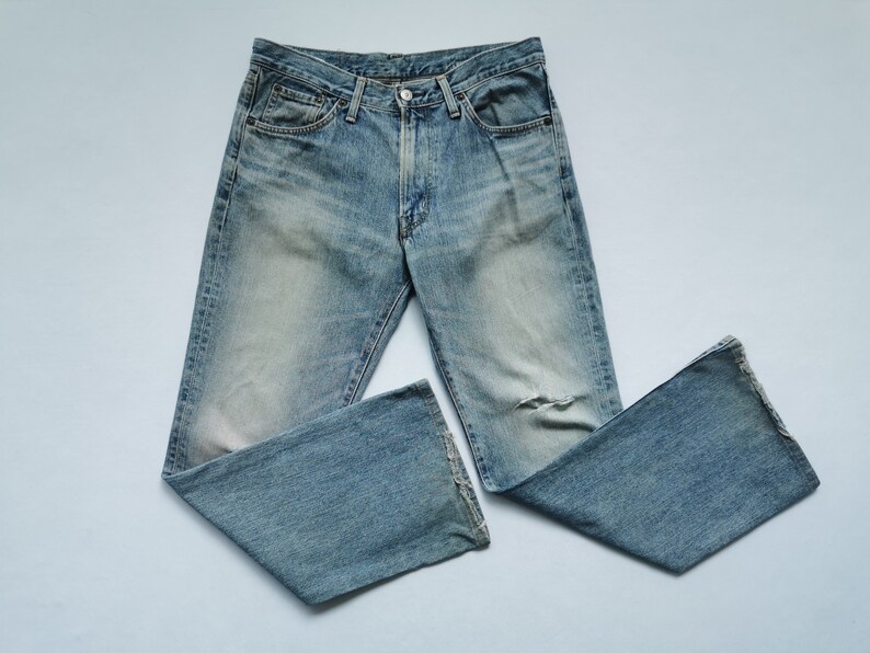 May include: Light blue denim jeans with a faded wash and a distressed knee. The jeans have a classic five-pocket design, a button and zip closure, and a straight leg. The jeans are laid flat on a light blue surface.