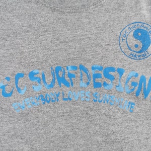May include: Gray t-shirt with blue text that reads "TEC SURF DESIGNS" and "EVERYBODY LOVES SUNSHINE". A blue yin-yang symbol is in a circle with the words "TEC SURF Designs Hawaii".