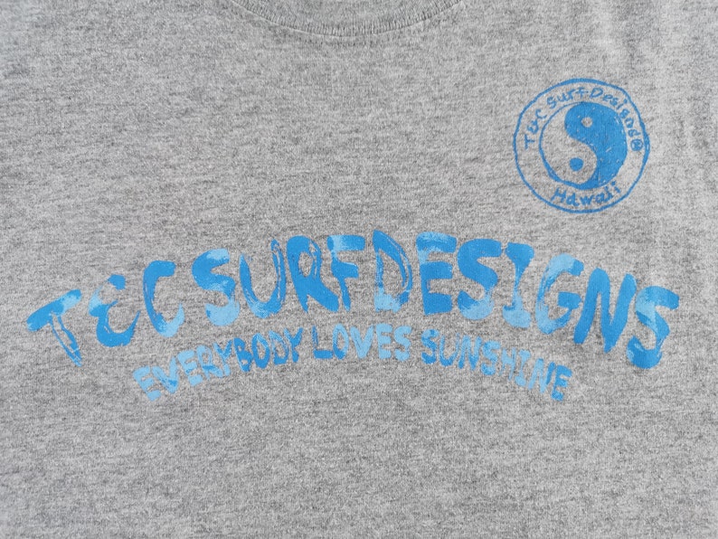 May include: Gray t-shirt with blue text that reads "TEC SURF DESIGNS" and "EVERYBODY LOVES SUNSHINE". A blue yin-yang symbol is in a circle with the words "TEC SURF Designs Hawaii".