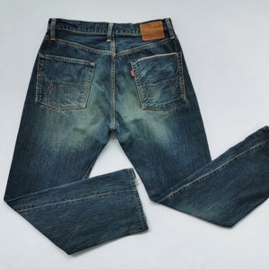 Pu&ograve; includere: Jeans in denim blu con lavaggio sbiadito e cuciture decorative sulle tasche posteriori. I jeans hanno un classico design a cinque tasche, una toppa in pelle e un'etichetta rossa. I jeans sono stesi su una superficie bianca.