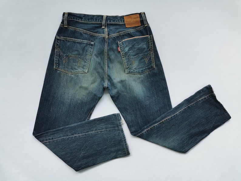 Pu&ograve; includere: Jeans in denim blu con lavaggio sbiadito e cuciture decorative sulle tasche posteriori. I jeans hanno un classico design a cinque tasche, una toppa in pelle e un'etichetta rossa. I jeans sono stesi su una superficie bianca.