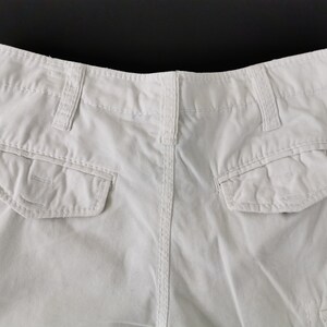 May include: A close-up view of the rear of white cargo shorts. The shorts feature two large pockets, belt loops, and a button fastening. The material looks to be a robust cotton or blend.