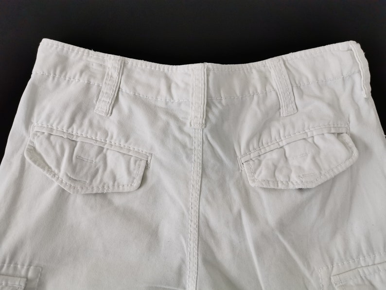 May include: A close-up view of the rear of white cargo shorts. The shorts feature two large pockets, belt loops, and a button fastening. The material looks to be a robust cotton or blend.